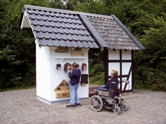 Eine Besucherin im Rollstuhl mit Elektroantrieb vor den Insektenhotels und Nistkästen