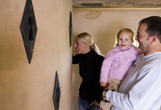 Eine Familie steht vor der Wand mit übergroßen Schlüssellöchern. Die Frau schaut durch eines hindurch