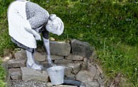 Eine Montage zeigt ein lebensgroßes schwarz-weiß-Bild einer älteren Dame, die mit einem Zinkeimer Wasser aus einem Brunnen holt.