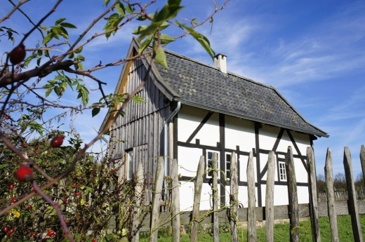 Kleinstwohnhaus aus Hilden mit sichtbarem Fachwerk und der verbretterten Wetterseite. Im Vordergrund der Gartenzaun und rote Hagebutten
