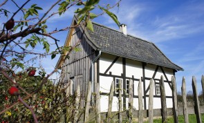 Kleinstwohnhaus aus Hilden mit sichtbarem Fachwerk und der verbretterten Wetterseite. Im Vordergrund der Gartenzaun und rote Hagebutten