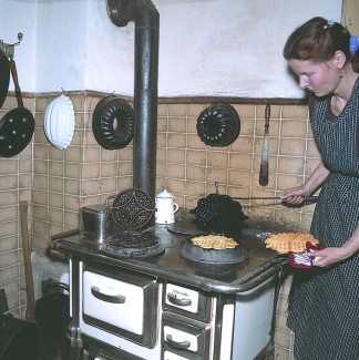 Die Hauswirtschafterin backt auf der 'Kochmaschine'  Waffeln mit einem gusseisernen Waffeleisen. Der Ofen wird mit Holz und Kohle befeuert.