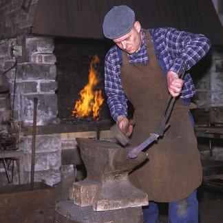 Der Schmied schmiedet das heiße Eisen mit einem Hammer am Amboss. Er trägt eine Lederschürze und im Hintergrund erkennt man das Schmiedefeuer