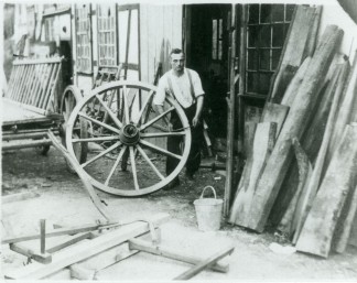 Historische Schwarzweiß-Aufnahme um 1935: Der Stellmacher Heinrich Bosbach vor seiner Werkstatt. Er präsentiert ein selbstgefertigtes hölzernes Wagenrad, das so groß ist wie er selbst.