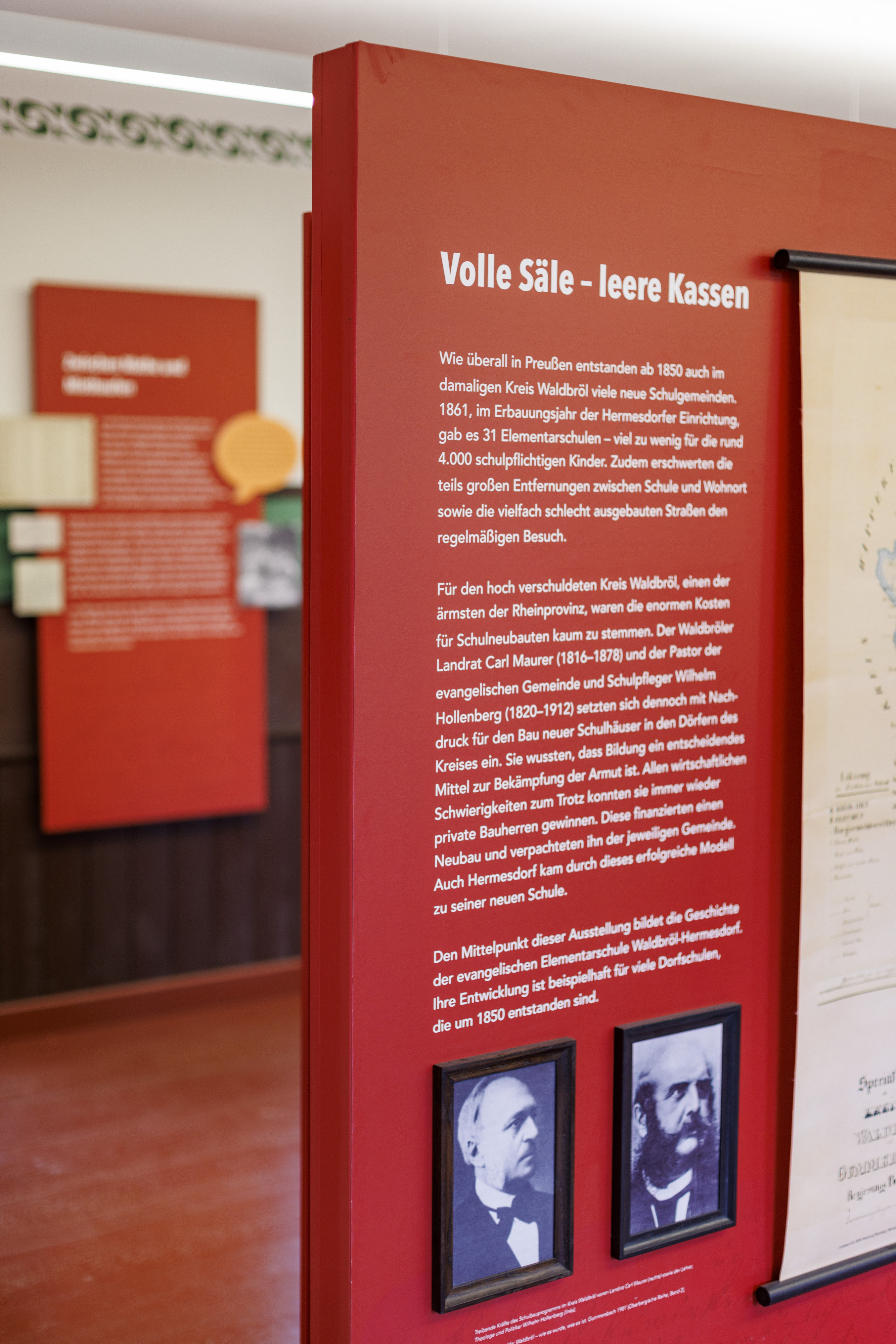 Auf einer roten Wand ist ein Ausstellungstext, Fotos und im Anschnitt eine große Landkarte zu sehen, die sich mit der historischen Entwicklung der Schulen beschäftigen.