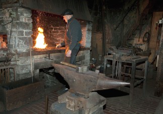 Der Schmied steht am Feuer. Im Vordergrund ist der große Amboss zu sehen.