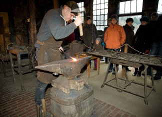 Der Schmied bearbeitet am Amboss Metall mit einem schweren Hammer. Im Hintergrund stehen Museumsbesucherinnen- und besucher.