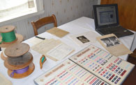 Auf einem Tisch liegen Rollen mit gewebten Bändern, Bücher mit Bandmustern und alte Fotos. Daneben steht ein Laptop.