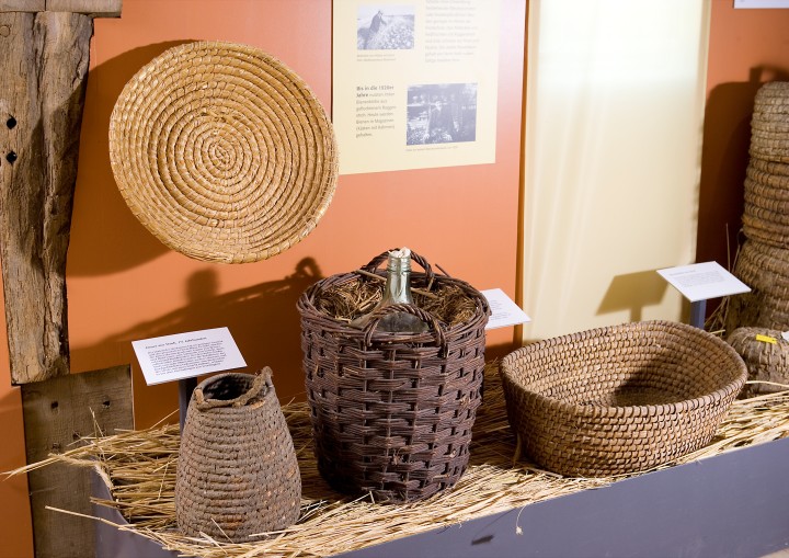 Ausstellungsvitrine mit kunstvoll gestalteten Körben aus Stroh.