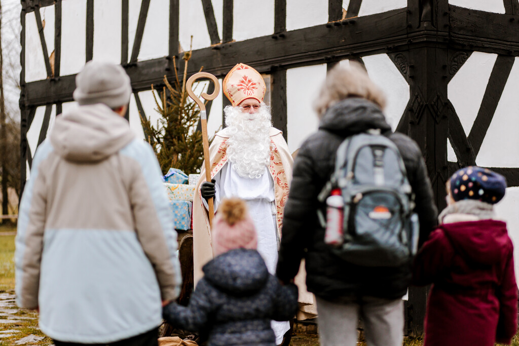 Zwei Erwachsene und zwei Kinder stehen vor einem als traditioneller Nikolaus verkleidetem Mann. Im Hintergrund sind ein Weihnachtsbaum, Geschenke und ein Fachwerkhaus zu sehen.