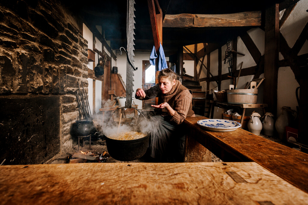 Eine Frau hockt in einerm de historischen Fachwerkhaus vor der Kochstelle. In dieser hängt ein großer Topf, in dem ein Eintopf kocht. Die Frau probiert den Eintopf mit einem Löffel.