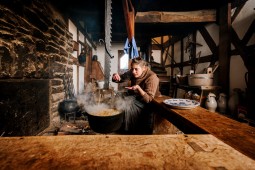 Eine Frau hockt in einerm de historischen Fachwerkhaus vor der Kochstelle. In dieser hängt ein großer Topf, in dem ein Eintopf kocht. Die Frau probiert den Eintopf mit einem Löffel.